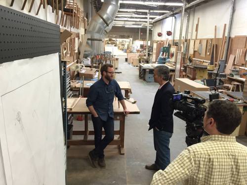 Two men converse in a woodworking shop, surrounded by tools and wooden materials, with a camera crew filming.