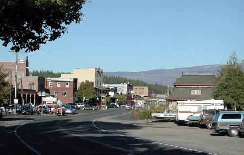 A small town street lined with shops and parked cars, with mountains in the background under a clear blue sky.