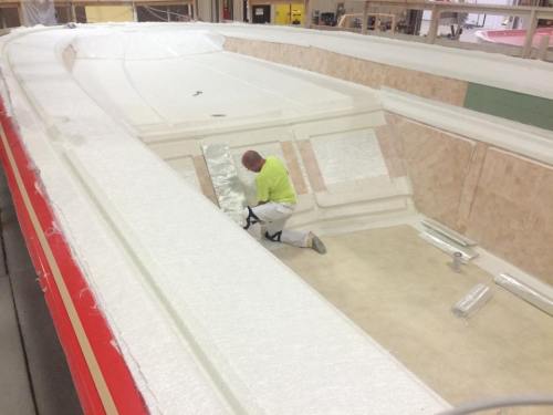 A worker in a bright shirt is applying material inside a boat hull under construction.