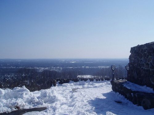A snowy landscape viewed from a high vantage point, with a stone wall and clear blue sky in the background.