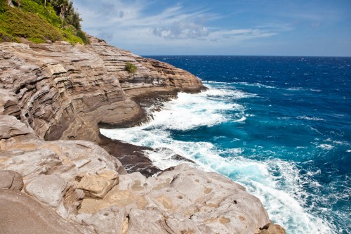 Rocky coastline with waves crashing against the shore under a blue sky with scattered clouds.