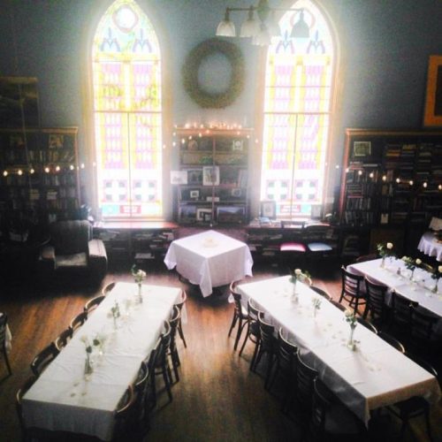 A cozy library with stained glass windows, featuring tables set for an event, adorned with flowers and soft lighting.
