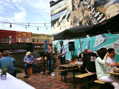 A lively outdoor scene with musicians performing near a colorful mural, while diners enjoy food at picnic tables.