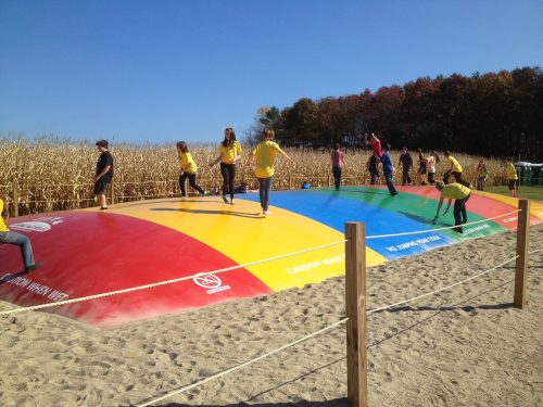 Children in yellow shirts play on a colorful inflatable bounce pad at a farm, surrounded by cornfields and blue skies.