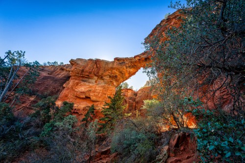 A natural rock arch surrounded by vibrant red cliffs and green foliage under a clear blue sky.