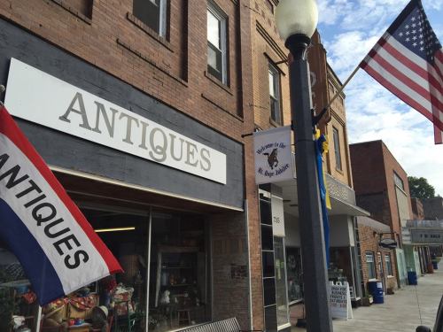 A storefront labeled "ANTIQUE" with flags and a classic brick building in a small town setting.