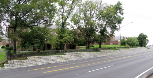 A view of a road lined with a stone wall and trees, with buildings visible in the background.