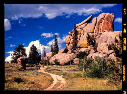 A scenic trail leads through grassy terrain, flanked by large rock formations and trees under a blue sky with clouds.