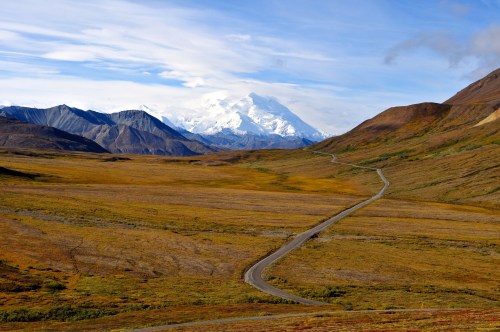 A winding road through a vast, colorful landscape with mountains and a snow-capped peak in the distance.
