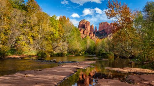 A serene river flows through a vibrant landscape of trees, with red rock formations and blue skies in the background.