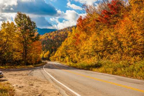 A winding road surrounded by vibrant autumn foliage and mountains under a partly cloudy sky.