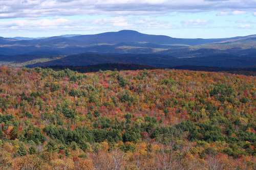 A panoramic view of colorful autumn foliage covering rolling hills and mountains under a partly cloudy sky.