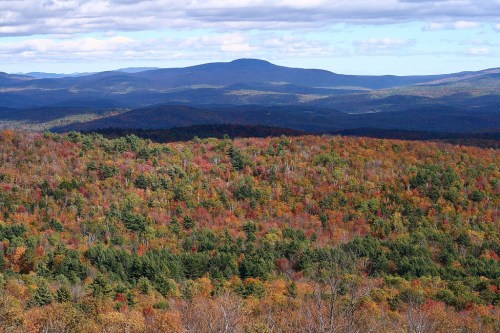 A panoramic view of colorful autumn foliage covering rolling hills and mountains under a partly cloudy sky.