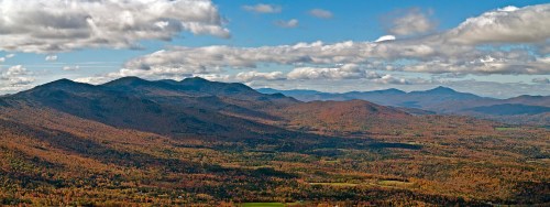 A panoramic view of colorful autumn mountains under a blue sky with scattered clouds.