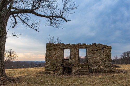 A weathered stone ruin with three windows stands in a grassy field under a cloudy sky, with a bare tree nearby.
