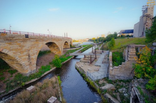 A scenic view of a river with a stone bridge, greenery, and remnants of old structures along the banks.