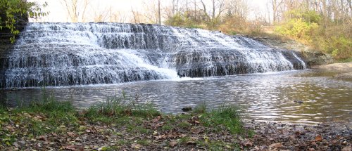 A serene waterfall cascading over rocks into a calm pool, surrounded by trees and autumn leaves.