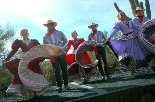 A group of dancers in traditional attire perform on stage, showcasing vibrant costumes against a desert backdrop.