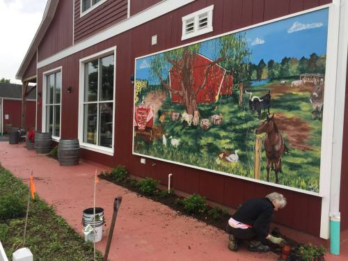 A person gardening in front of a colorful mural depicting a farm scene with animals and a red barn.