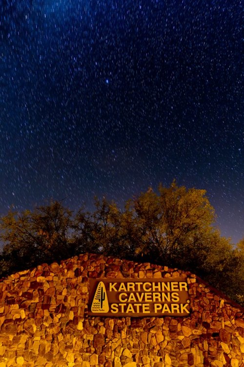 A starry night sky above the Kartchner Caverns State Park sign, surrounded by trees and rocky terrain.