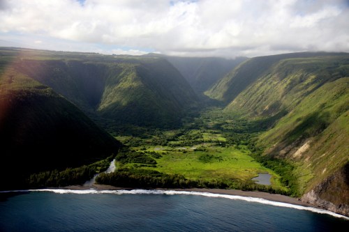 Aerial view of a lush green valley surrounded by steep cliffs, with a river and ocean at the base.