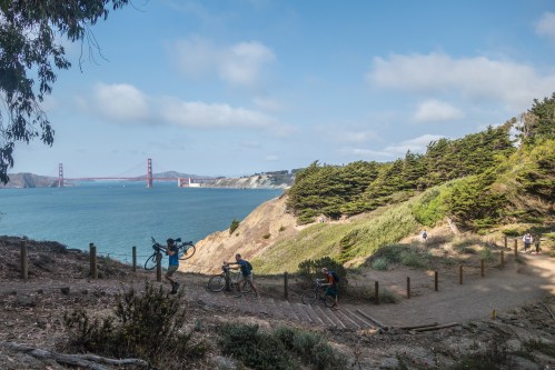 A scenic coastal path with cyclists, overlooking the ocean and the Golden Gate Bridge in the background.