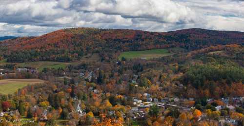 A scenic view of a small town surrounded by vibrant autumn foliage and rolling hills under a cloudy sky.