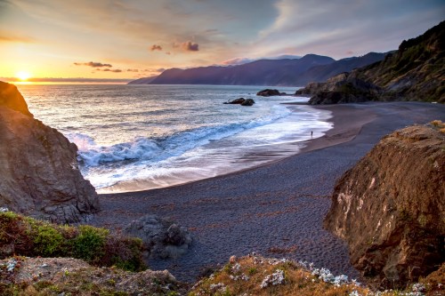 A serene beach at sunset, with gentle waves, rocky cliffs, and a person walking along the shore.