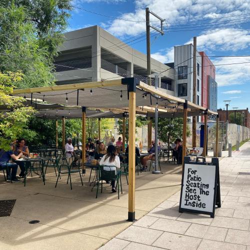 Outdoor patio seating area with tables and chairs, surrounded by greenery, and a sign directing to the host inside.