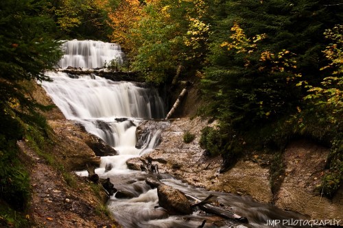 A serene waterfall cascades down rocky steps, surrounded by vibrant autumn foliage and lush greenery.