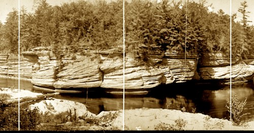 A sepia-toned landscape featuring layered rock formations beside a calm river, surrounded by dense trees.
