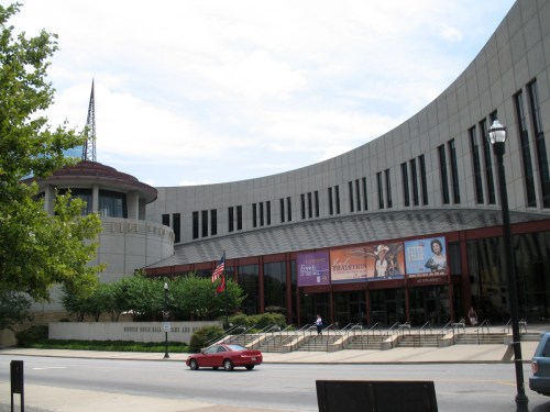 Exterior view of a modern building with curved architecture, featuring banners and greenery in front.