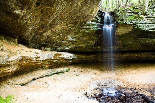 A serene waterfall cascades over rocky cliffs into a sandy area, surrounded by lush greenery and natural rock formations.