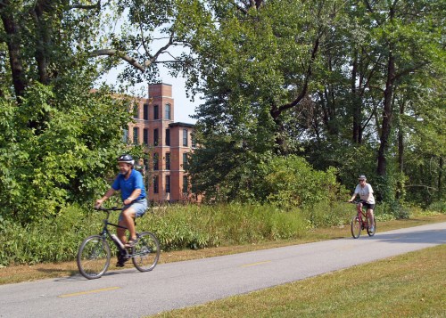 Two cyclists ride along a paved path surrounded by greenery, with a brick building visible in the background.