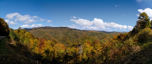 Panoramic view of rolling hills covered in vibrant autumn foliage under a clear blue sky with scattered clouds.