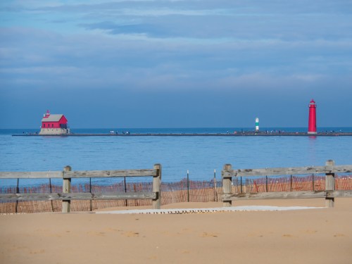 A sandy beach with a wooden fence, featuring a red lighthouse and a small building on a pier in calm waters.