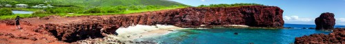 A panoramic view of a rocky coastline with red cliffs and clear blue water under a bright sky.