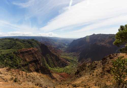 A panoramic view of a lush valley surrounded by steep, rocky cliffs under a blue sky with wispy clouds.