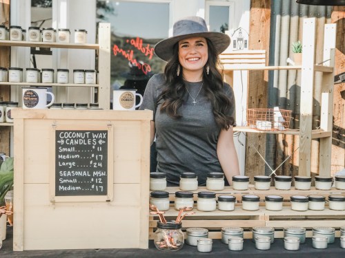 A smiling woman in a hat stands behind a wooden display of coconut wax candles, showcasing prices on a chalkboard.