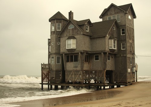 A large, unique wooden house on stilts by the beach, with waves crashing nearby under a cloudy sky.
