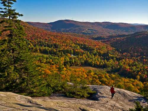 A hiker stands on a rocky outcrop, overlooking a vibrant autumn landscape filled with colorful trees and distant hills.