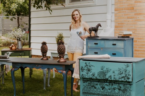 A woman stands among colorful furniture pieces in a garden, holding a paintbrush and smiling.