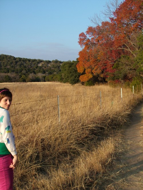 A person in a colorful sweater stands by a grassy path, with autumn trees and a fence in the background.