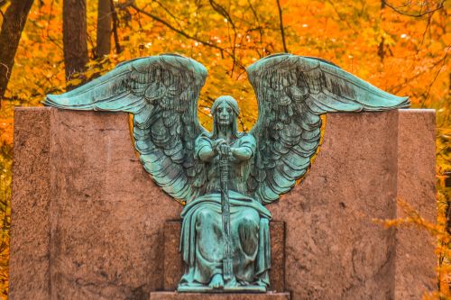 A bronze angel statue with large wings sits on a stone pedestal, surrounded by vibrant autumn foliage.