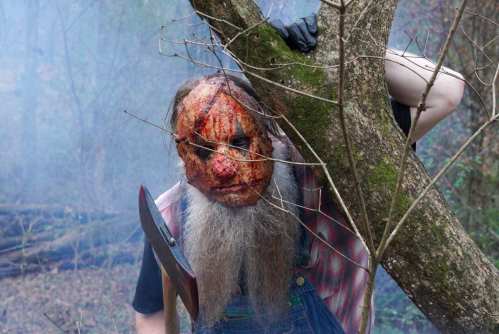 A figure with a gruesome mask and axe peeks from behind a tree in a foggy, eerie forest setting.
