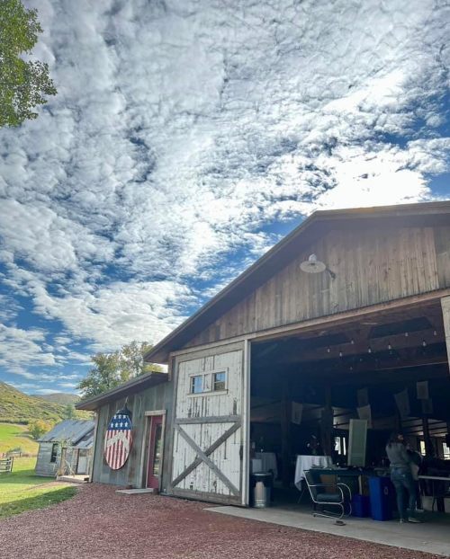 A rustic barn with an American flag mural, under a sky filled with fluffy clouds. Green hills are visible in the background.