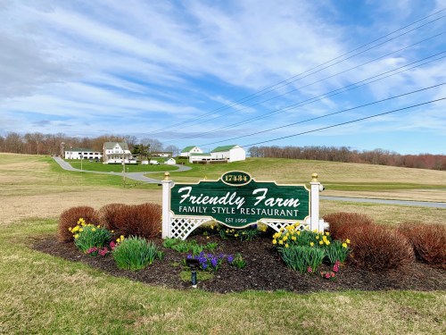 Sign for Friendly Farm Family Style Restaurant, surrounded by colorful flowers, with a scenic landscape in the background.