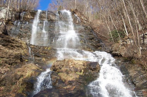 A cascading waterfall flows down rocky cliffs, surrounded by trees and a clear blue sky. A faint rainbow appears in the mist.