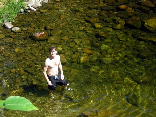 A young man stands in a clear, shallow stream surrounded by rocks and greenery, enjoying a sunny day.