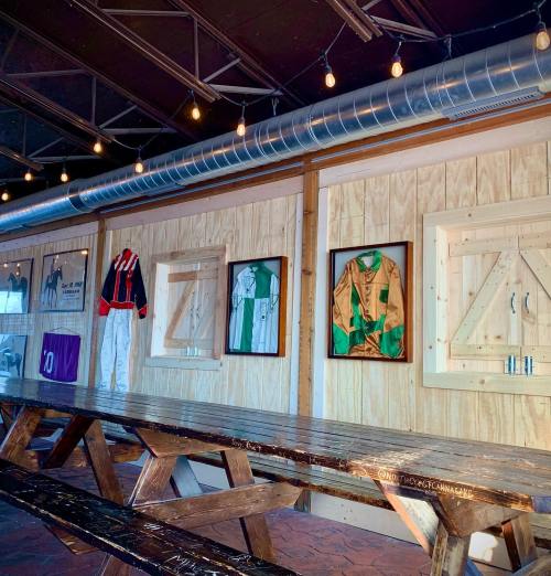 Interior of a rustic space featuring framed jockey outfits on wooden walls and a long picnic-style table.
